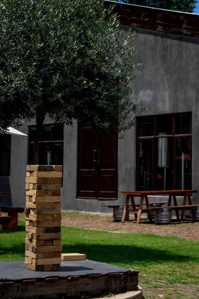 A vibrant outdoor scene featuring a large Jenga game set under a tree in a backyard on a sunny day.