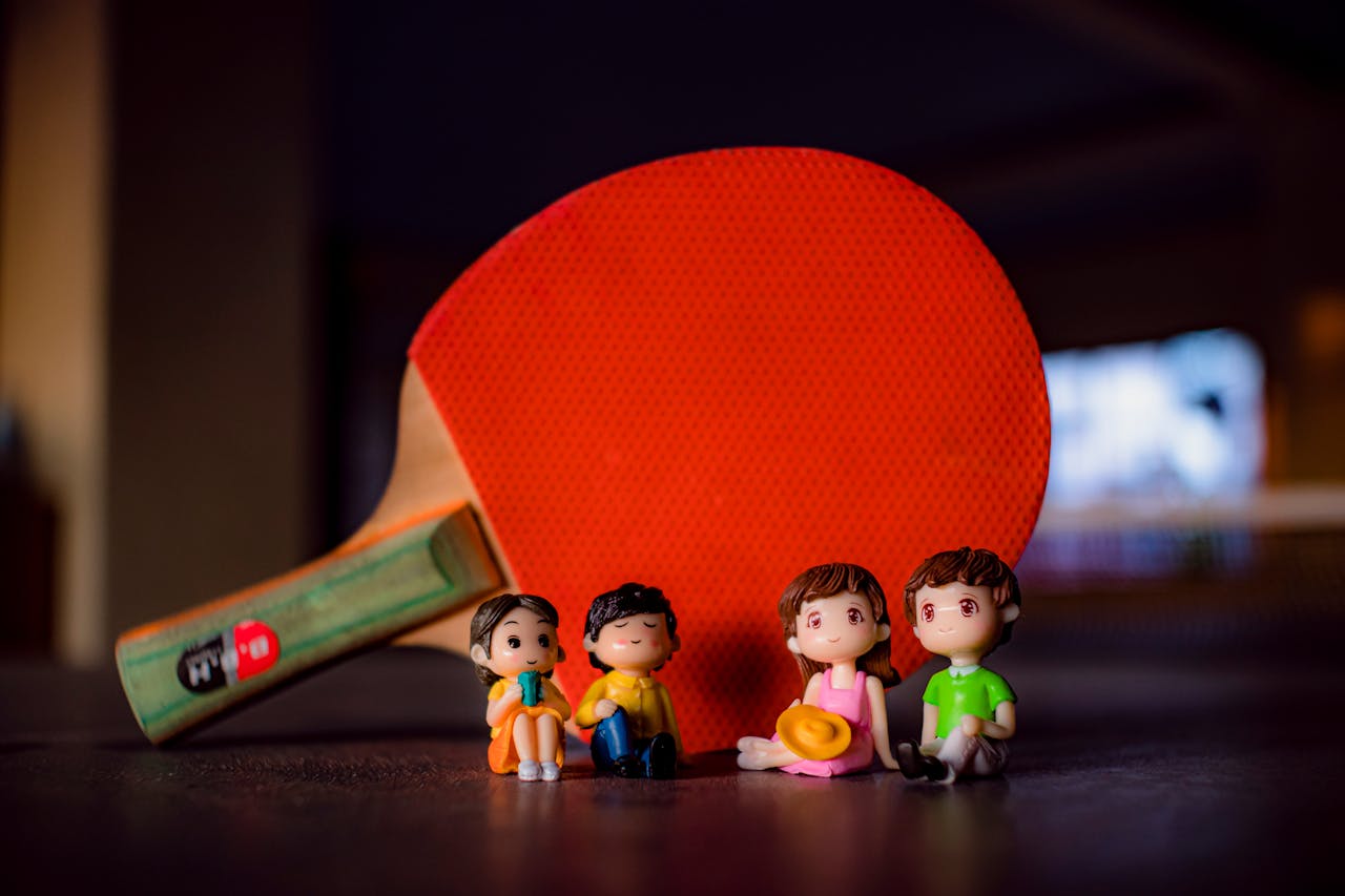 Small colorful figurines of couples placed on table near professional red table tennis racket in light room on blurred background